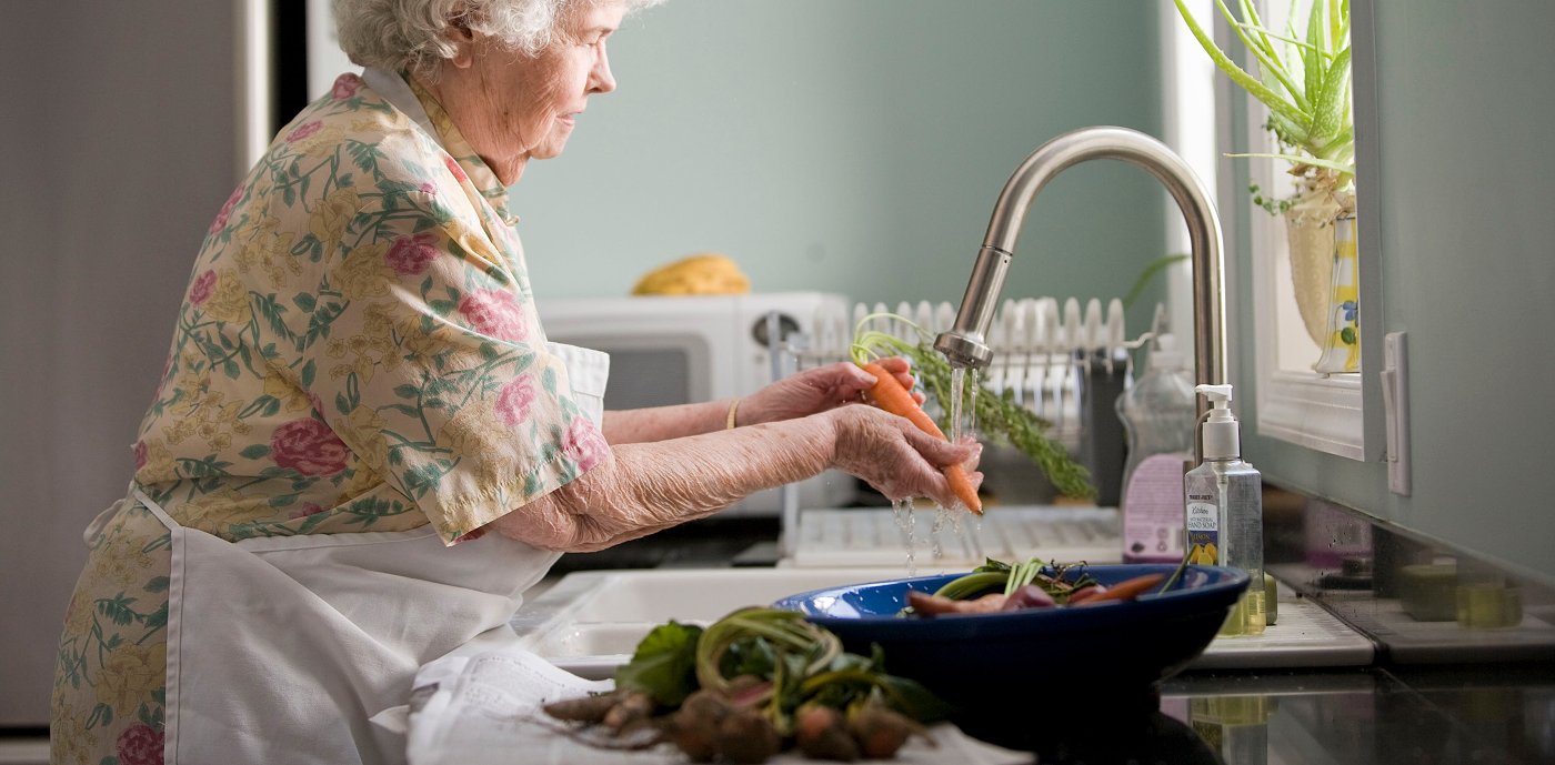 Eldre kvinne vasker grønnsaker i kjøkkenvask