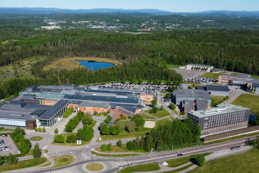  Luftfoto av moderne campus eller industrikompleks med solcellepaneler, omgitt av skoger og fjell, med en liten innsjø i nærheten.