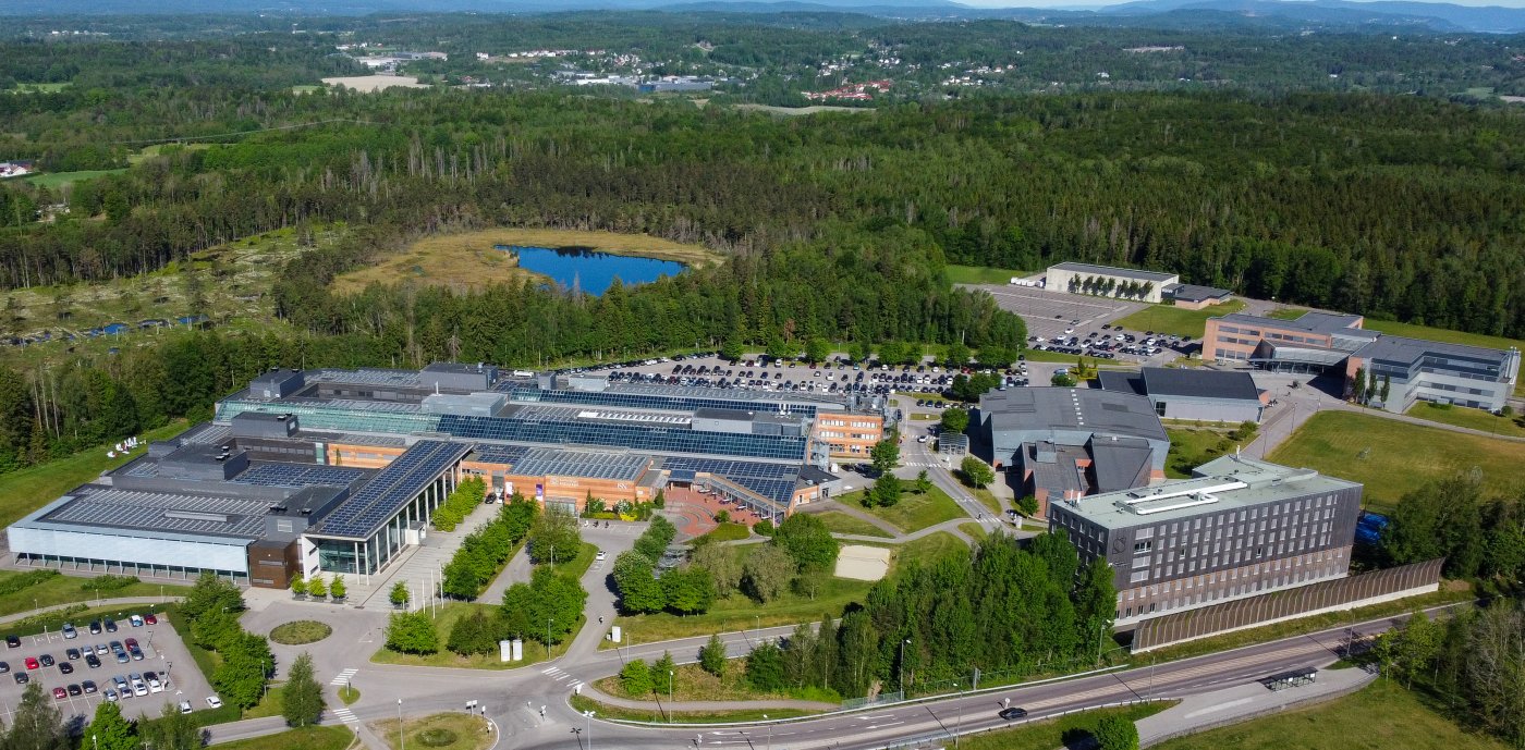  Luftfoto av moderne campus eller industrikompleks med solcellepaneler, omgitt av skoger og fjell, med en liten innsjø i nærheten.